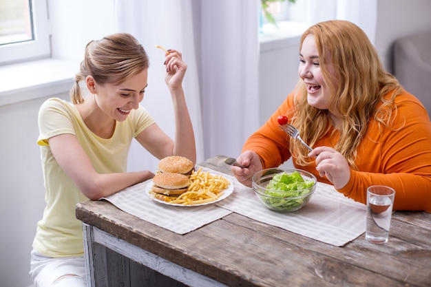 Amazing day. Laughing slim woman eating fast food and talking with her fat friend eating a salad