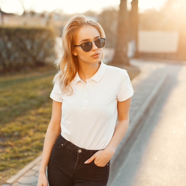 Amazing fashionable young hipster woman in stylish summer clothes is standing and posing at sunset of summer. Attractive girl. American style clothing.