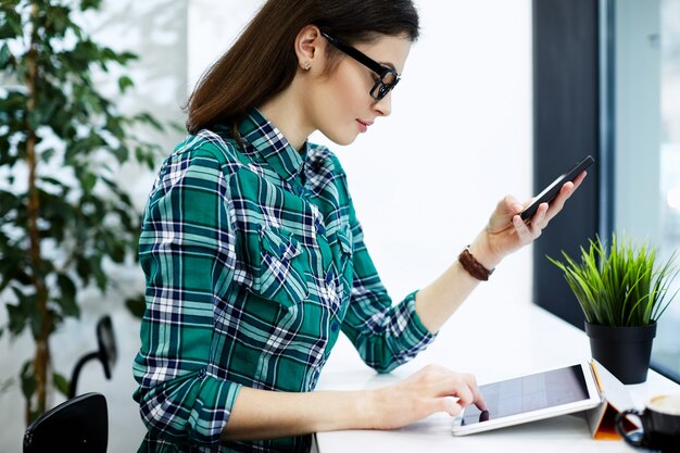 Amazing girl with black hair wearing shirt and eyeglasses sitting in cafe with tablet and cup of coffee, holding mobile phone, freelance concept.