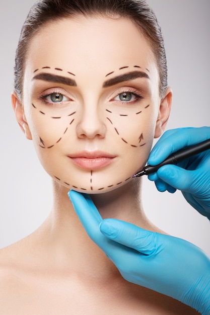 Amazing girl with dark eyebrows at studio background, doctor's hands wearing blue gloves drawing perforation lines on face, plastic surgery concept.