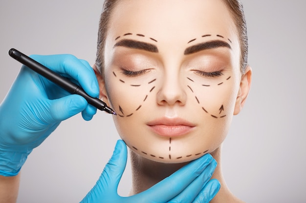 Incredibile ragazza con le sopracciglia scure al fondo dello studio, le mani del medico indossando guanti blu disegnando linee di perforazione sul viso, concetto di chirurgia plastica.