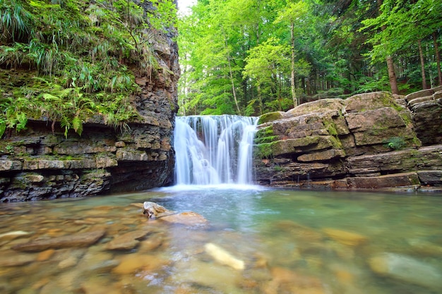 Amazing landscape of beautiful waterfall on mountain river with white foamy water falling down from rocky cliff in summer rainforest.