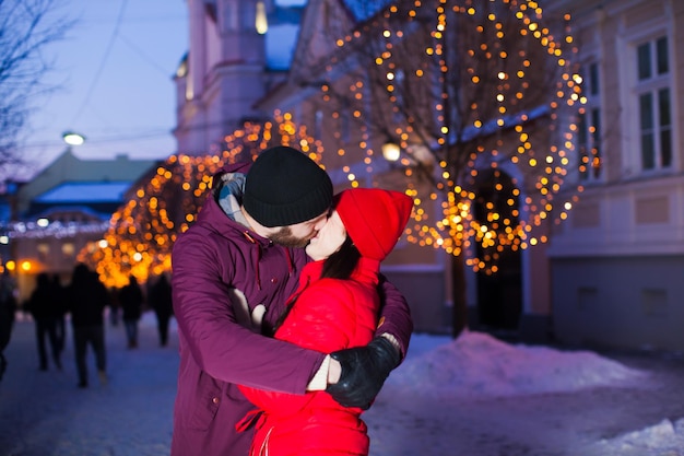 Amazing romantic couple embracing and kissing on the Christmas city background. Handsome man holding tightly his young wife, bright lights on the trees of old city of Uzhgorod on the background