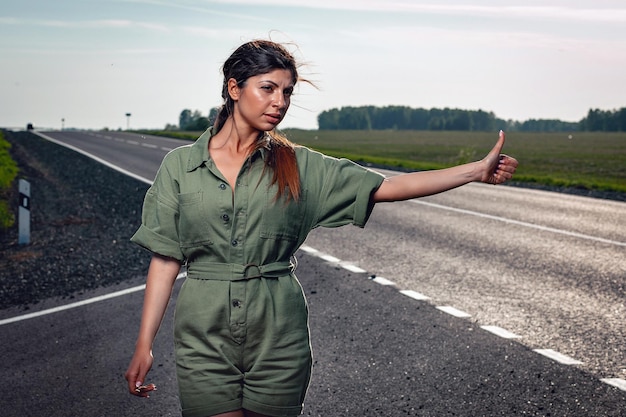 タイトなオーバーオールの驚くべき薄い女性が道路でヒッチハイクしている、肖像画