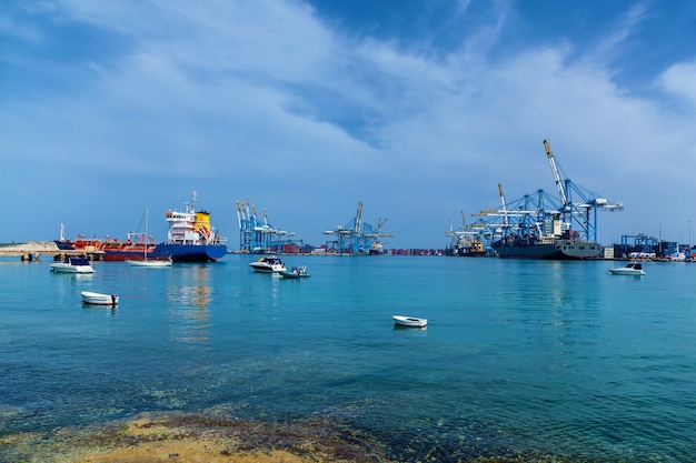 An amazing view of an industrial port with many cargo ships near construction cranes on the sea on the background of a blue sky in Malta.