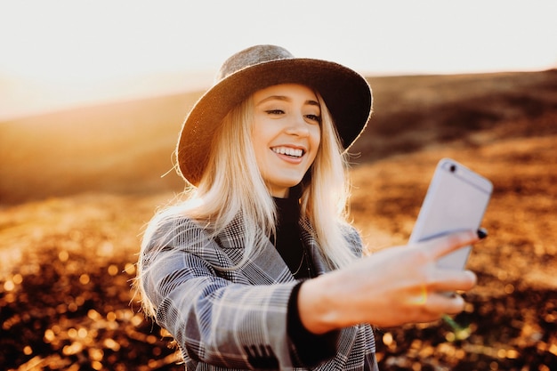 Amazing woman with white hair dressed in suit and wearing hat doing a selfie smiling against beautiful landscape at the sunset.