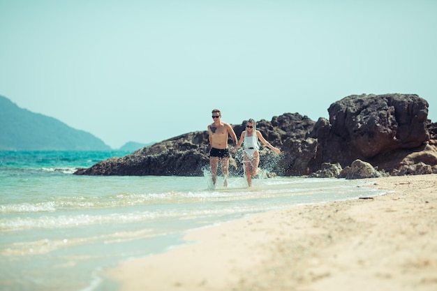 驚くべき若いカップルは、水泳パンツの男と彼のガールフレンドと手をつないでビーチを走ります