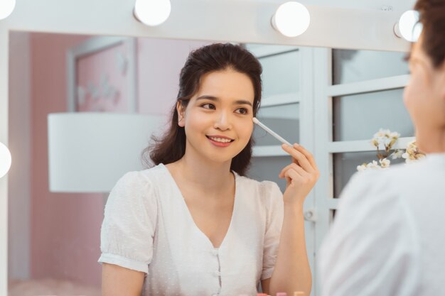 Amazing young woman doing her makeup in front of mirror. Portrait of beautiful girl near cosmetic table