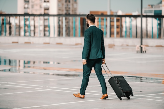 Ambitieuze blanke bebaarde zakenman in formele slijtage met bagage en wandelen op parkeerplaats. bedrijfsconcept reis. ruggen draaiden zich om. succes komt voor wanneer je dromen groter worden dan je excuses.