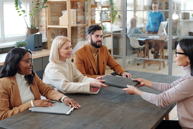 Ambitieuze jonge Aziatische vrouw met een bril die aan tafel zit voor recruiters en hen haar portfolio geeft tijdens een sollicitatiegesprek