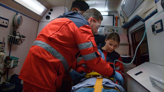 写真 救急車の医師が救急車で被害者の命を救う 医療チームが男をストレッチャーで固定する 多民族の救急隊員が病院の途中で負傷した患者に酸素マスクをかける 医療コンセプト