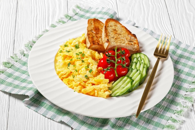 Colazione americana di uova strapazzate con cetriolo affettato, pomodori, pane tostato su un piatto bianco con forchetta dorata su un tavolo strutturato in legno