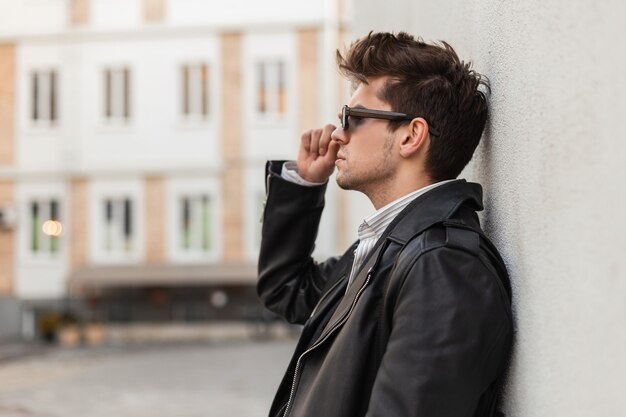 Брутальный американский молодой человек со стильной прической в модной негабаритной черной кожаной куртке поправляет винтажные темные очки у серой стены на улице. Городской парень позирует в городе на улице