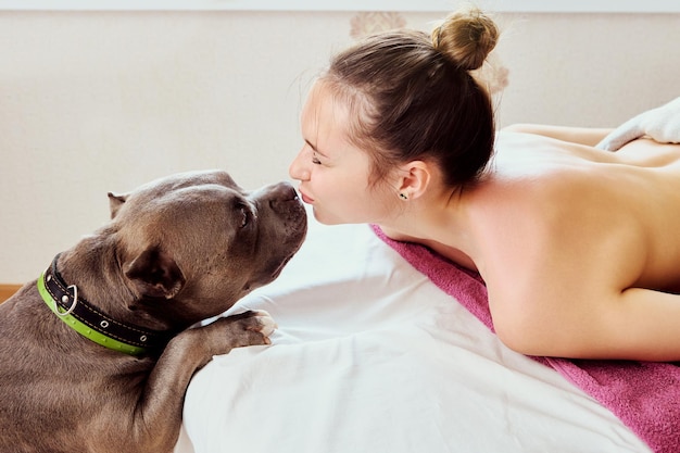 Photo an american bully dog kisses young woman who is lying on massage table in a massage parlor