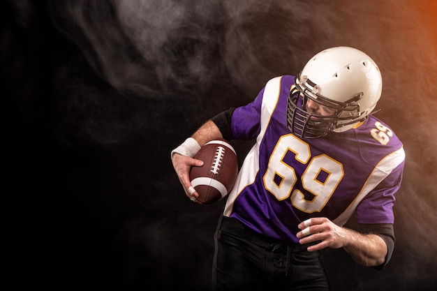 American football player holding ball in his hands in smoke\
black background copy space the concept of american football\
motivation copy space