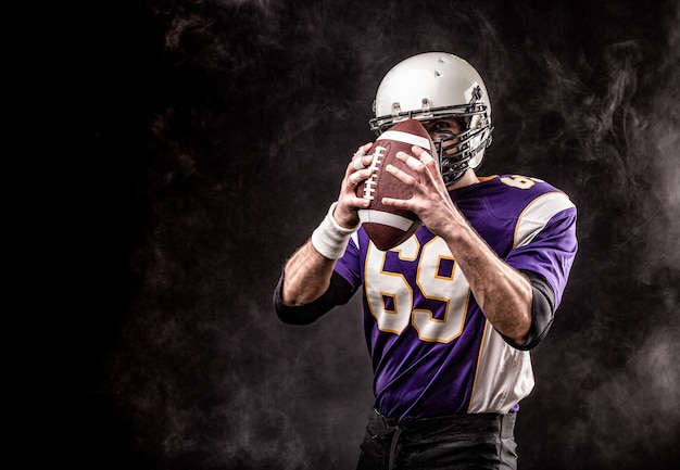 American football player holding ball in his hands in smoke. Black wall, copy space. The concept of American football, motivation, copy space