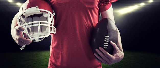 American football player holding a helmet and ball against rugby stadium