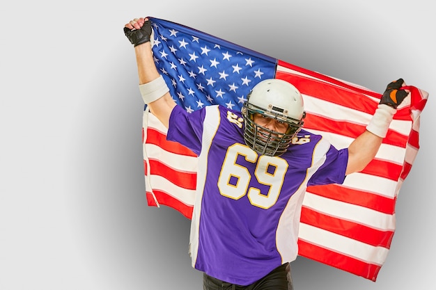 Giocatore di football americano con uniforme e bandiera americana orgoglioso del suo paese, su un muro bianco