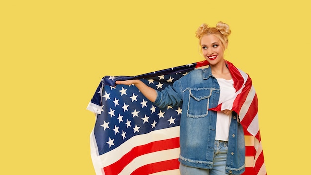 American girl. Happy young woman in denim clothes holding USA flag isolated on yellow background. 