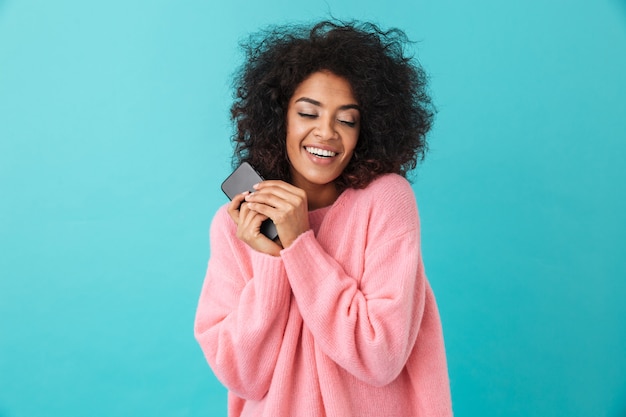 American happy woman in pink shirt smiling and holding black mobile phone in hands, isolated over blue wall