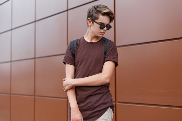American hipster young man with a fashionable hairstyle in sunglasses in fashionable clothes with a black backpack poses near a wall