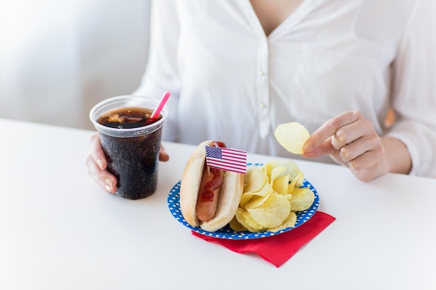 アメリカの独立記念日、お祝い、愛国心、祝日のコンセプト – 7月4日のホームパーティーで、ホットドッグとコカ・コーラをプラスチックカップに入れてポテトチップスを食べる女性の接写