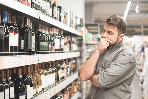 american man at jeans jacket and black beret holding basket and looking on bottle of wine, shopping at supermarket.