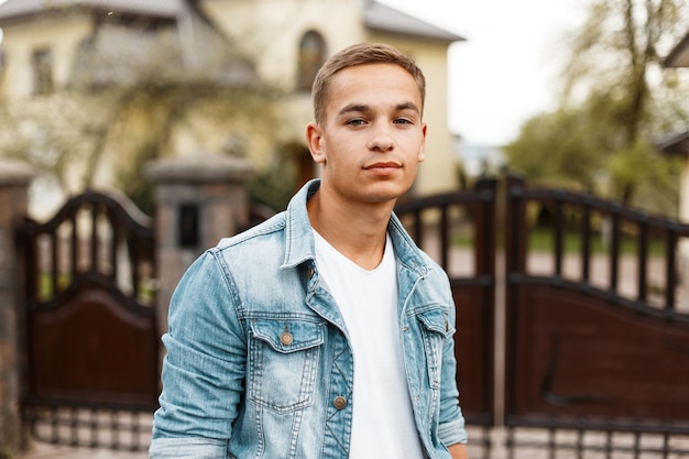 American modern attractive guy with a hairstyle in a stylish white T-shirt in a blue denim jacket stands near the old wrought iron gates near the house