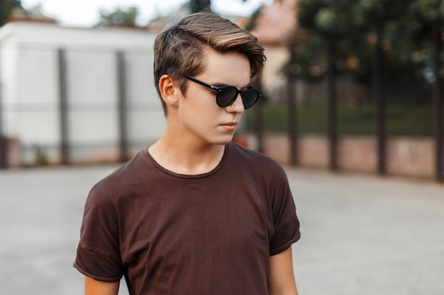 American modern young hipster man in stylish sunglasses in summer t-shirt is standing at the stadium on a warm summer day.