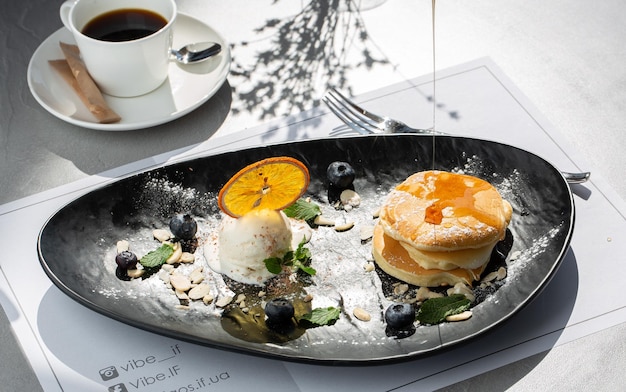 American pancakes with ice cream scoop, berries and maple syrup on black plate isolated with coffee. cafe menu