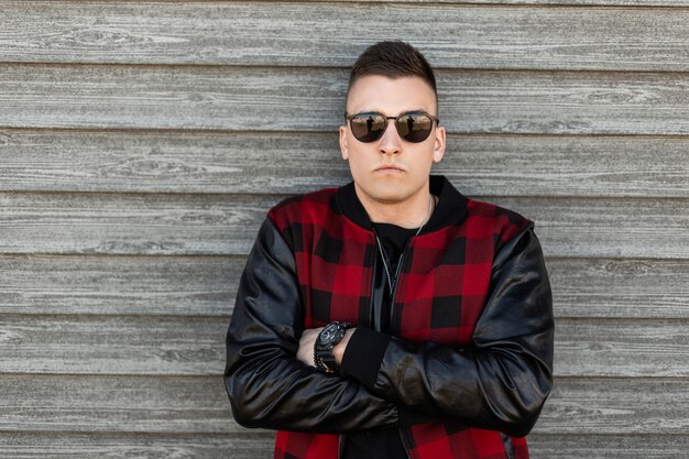 American serious young hipster man in black sunglasses in a vintage red checkered jacket in a black t-shirt with a stylish hairstyle near a wooden wall outdoors