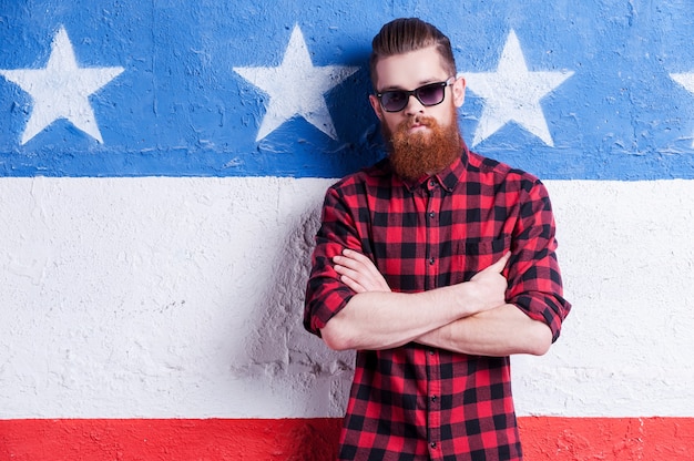 American style. Handsome young bearded man in sunglasses keeping arms crossed and looking at camera while standing against American flag