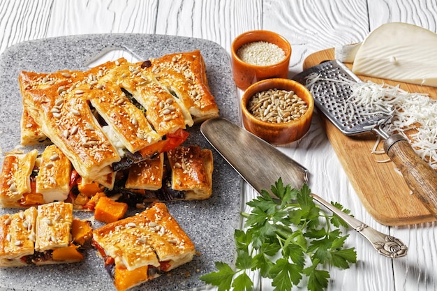 American vegetable puff pastry pie with goat cheese topped with sunflower and sesame seeds filled with roasted vegetables served on a white wooden background, top view, close-up