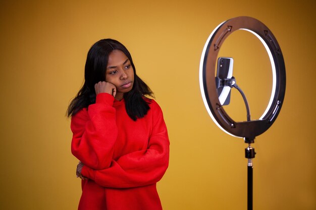 American vlogger girl in a red hoodie with a lighted ring lamp and phone resting head on hand