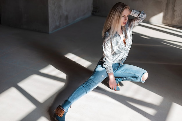 American young blonde woman in stylish shirt in vintage ripped jeans in trendy cowboy boots posing sitting indoors with sunlight