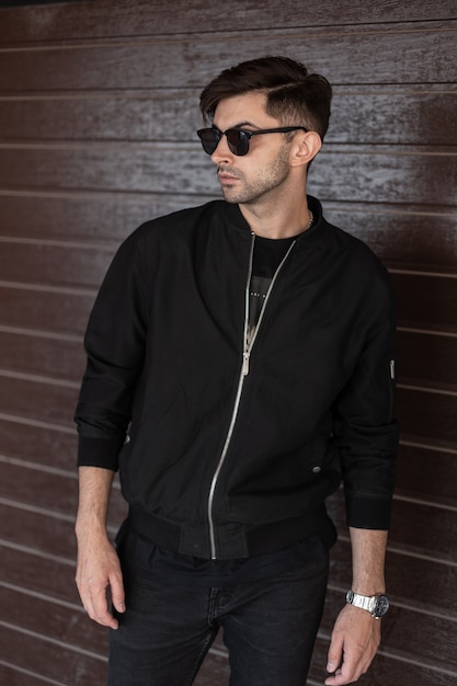 American young man in fashionable black clothes in stylish sunglasses with trendy hairstyle stands near a vintage brown wooden wall in the city. Handsome urban guy. Street fashion. Menswear.