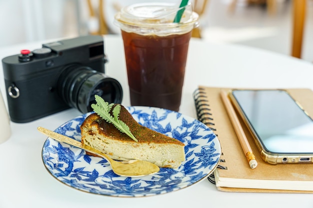 Americano iced black coffee in cup mug burnt face cheesecake placed and camera smartphone in notebook on wood desk on top view As breakfast In a coffee shop at the cafeduring business work concept
