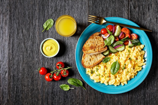 Amerikaans ontbijt van roerei met komkommer, spinazie-tomatensalade, geroosterd brood en een glas verse jus d'orange op een blauw bord op een donkere houten rustieke tafel, plat gelegd