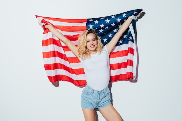 Amerikaanse blonde vrouw die de vlag van de VS houdt die over een witte muur wordt geïsoleerd