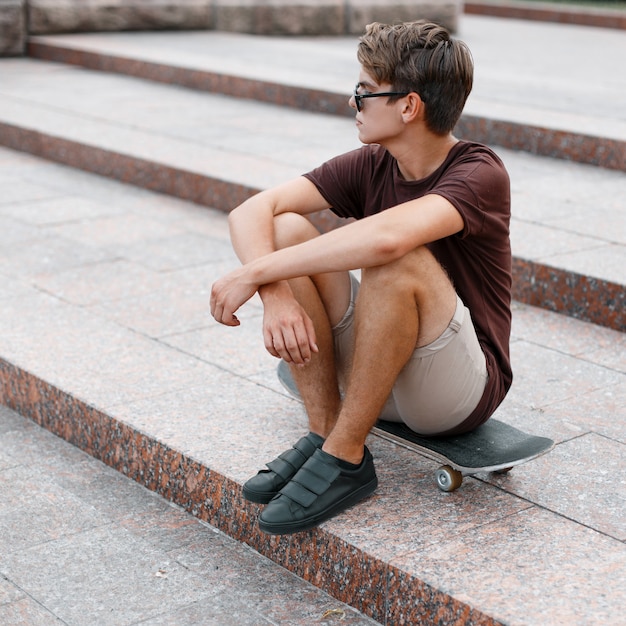 Amerikaanse coole hipster jongeman met kapsel in stijlvolle zonnebril in zomer t-shirt in korte broek in sneakers zittend op een skateboard