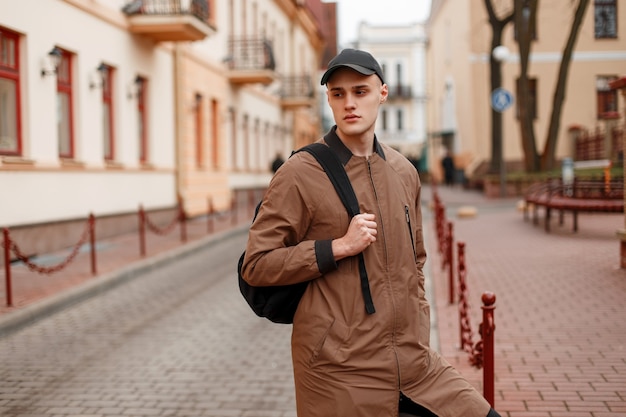 Amerikaanse modieuze knappe jongeman in een stijlvolle beige lange jas in een stijlvolle pet met een zwarte rugzak loopt door de stad op een lentedag