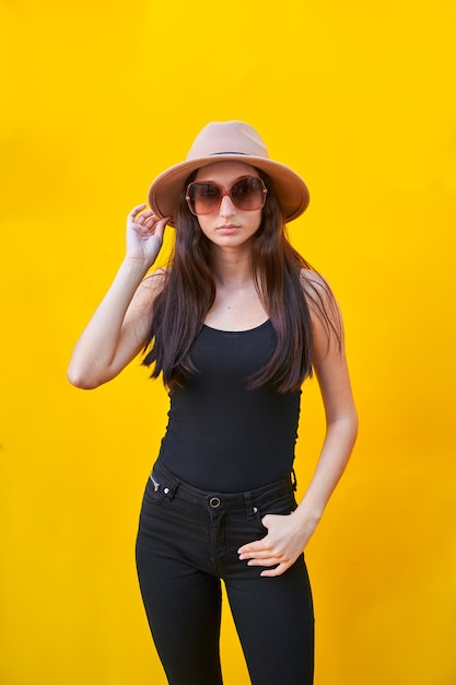 Amerikaanse platte jonge blanke vrouw met zonnebril voor lang haar, beige hoed, mouwloos onderhemd en zwarte broek, die de hoed met één hand vasthoudt