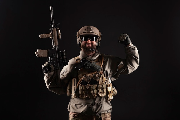 Amerikaanse soldaat in een militair pak met een geweer glimlacht en toont kracht tegen een donkere achtergrond