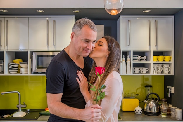 Amorous couple celebrating Valentine day at home in the kitchen. Romantic couple with rose while standing in kitchen