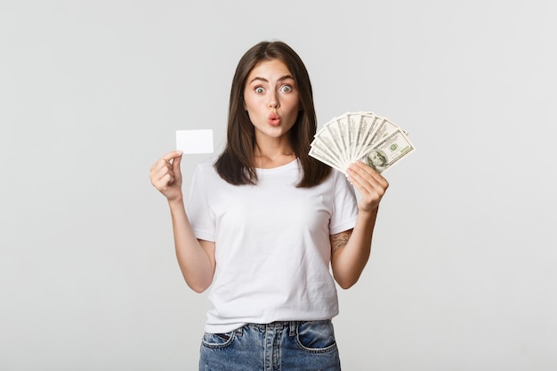 Amused brunette girl looking excited, holding money and credit card, standing white.