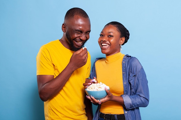 Amused couple watching comedy film on tv and having snack over blue background. Cheerful people laughing at funny movie, eating popcorn and enjoying millennial activity together.