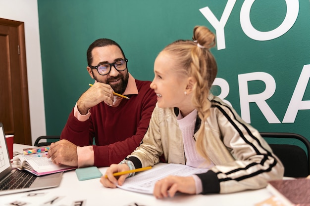 Amused Darkhaired bearded adult teacher wearing eyewear and his pupil looking amused while working at the lesson