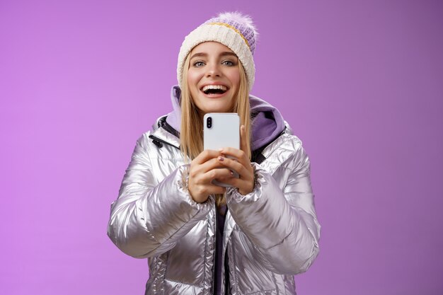Забавная возбужденная привлекательная белокурая подруга, держащая смартфон, записывает видео, шаг парня на сноуборде, впервые запечатлевая воспоминания, мобильная камера, счастливо стоящая на фиолетовом фоне.