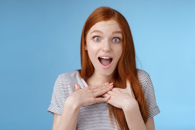 Amused happy grateful european redhead woman gasping drop jaw joyfully press palms chest thankful staring surprised incredible cool gift receive awesome proposal standing blue background.