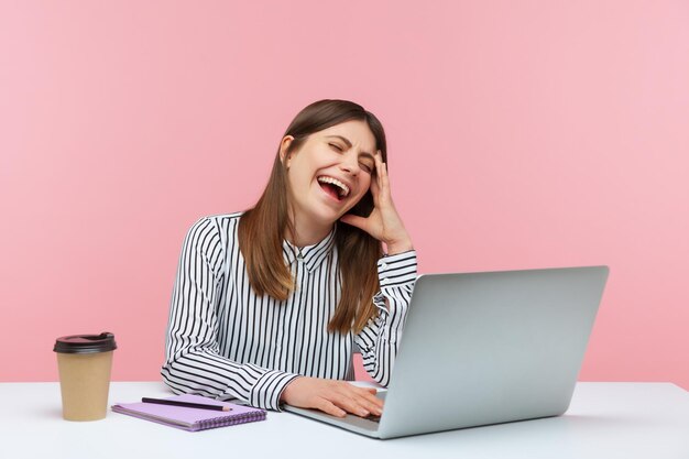 Amused happy woman office worker laughing out loud sitting at workplace enjoying funny joke on laptop having break at home office Indoor studio shot isolated on pink background